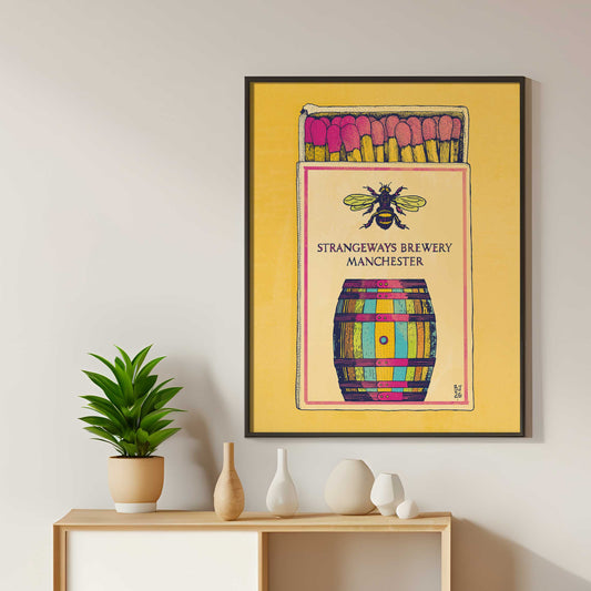 A framed art print featuring a colorful matchbox-style illustration labeled 'Strangeways Brewery Manchester' with a bee and barrel.