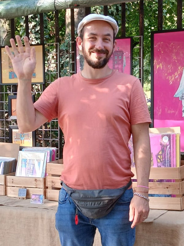 A man wearing a salmon-pink t-shirt, blue jeans, and a white cap stands smiling and waving in front of a market stall. The stall has a beige tablecloth, stacked book boxes, and colorful art prints hanging on a fence in the background.