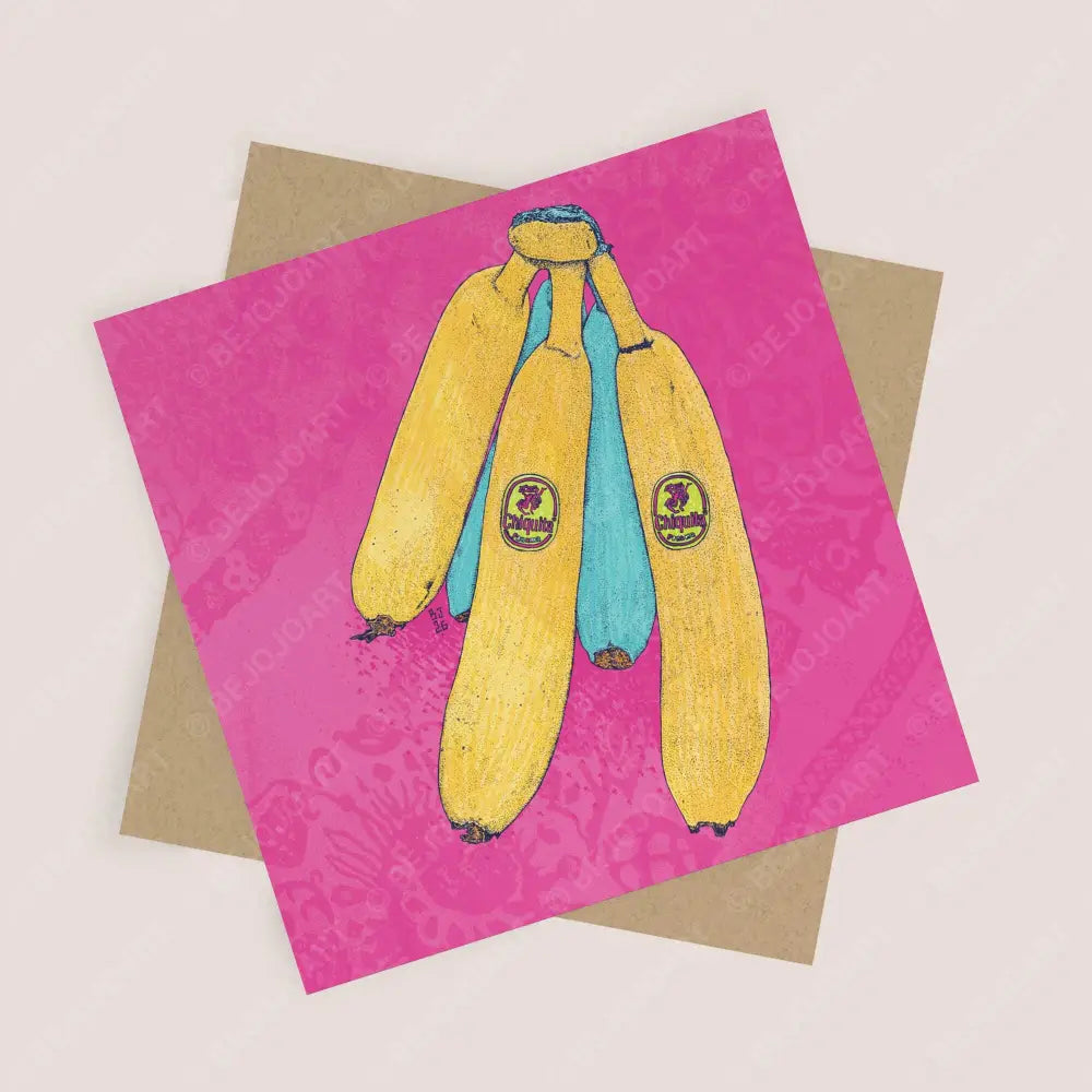 A square greeting card featuring a hand-drawn illustration of a bunch of yellow and blue bananas on a bright pink patterned background, shown with a brown kraft envelope on a light beige surface.