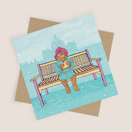 Christmas card showing a bear sitting on a bench in the snow with a city landmark behind